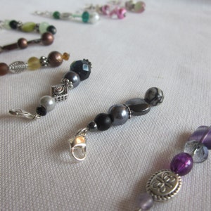May include: Close-up of several beaded jewelry charms with silver-tone clasps. The charms feature a variety of beads in colors like brown, black, gray, purple, and green. Some charms have decorative metal accents.