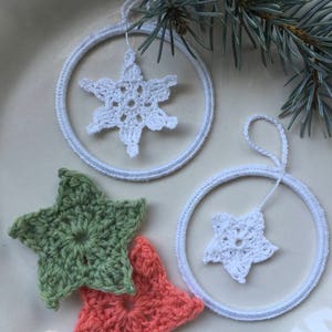 Könnte beinhalten: Drei gehäkelte Ornamente, zwei weiße Sterne in weißen Ringen und ein grüner Stern. Die Ornamente befinden sich auf einer weißen Oberfläche.