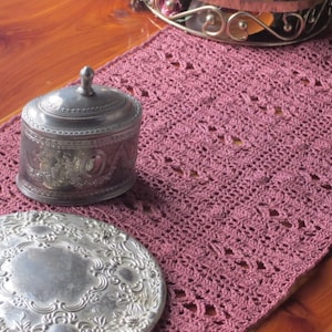 May include: A silver oval-shaped trinket box with an ornate design sits on a pink crocheted table runner. A silver tray with a similar design is in the foreground.