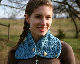 May include: A close-up of a light blue crocheted cowl with gold buttons. The cowl is worn around the neck and has a textured, open weave design. The background is blurred, with a person wearing the cowl.