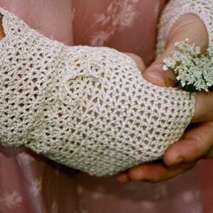 May include: A pair of white crocheted gloves with a delicate lace pattern. The gloves are worn by a person holding a bouquet of white flowers.