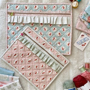 May include: Two quilted zippered pouches featuring a cherry pattern. The larger pouch is light blue with a beige ruffle, and the smaller pouch is pink with a light green ruffle. Both have matching fabric binding and metal zippers.