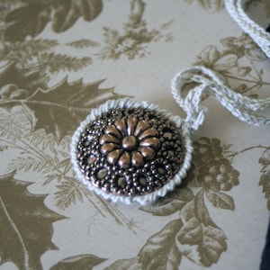 May include: A white crocheted necklace with a silver and copper pendant featuring a floral design. The pendant is round and has a raised center with a flower shape.