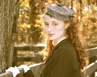 May include: A woman with long, curly red hair wears a brown coat and a gray and black hat with a veil. She is standing in front of a wooden fence with a tree in the background.