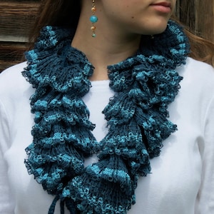 May include: A teal blue crocheted ruffle scarf with a layered design. The scarf is worn around the neck of a person.