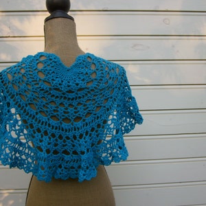 May include: A turquoise crocheted shawl with a scalloped edge. The shawl is displayed on a white wooden background.