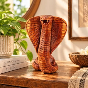 May include: A brown, carved wooden cobra figurine in a striking pose with its hood flared and mouth open, revealing fangs. The sculpture features detailed scale textures across its body and hood, showcasing a natural wood grain finish throughout.