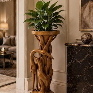 May include: A wooden plant stand with a carved design featuring two birds and foliage. The stand holds a potted plant with large green leaves. The stand is made of light brown wood and is placed on a marble floor.