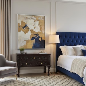Op de afbeelding: Een slaapkamer met een marineblauw gecapitonneerd hoofdbord, een donkere houten dressoir met gesneden details en een groot abstract schilderij met goud, wit en marineblauw. Een patroon tapijt en een gouden vloerlamp maken de inrichting compleet.