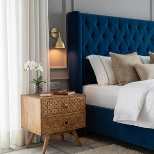 Könnte beinhalten: Schlafzimmer-Interieur mit einem marineblauen, getufteten Kopfteil, einem Nachttisch aus Holz mit geschnitzten Details und einer Wandleuchte aus Messing. Der Nachttisch hat zwei Schubladen mit Messinggriffen. Weiße Bettwäsche und gemusterte Kissen.