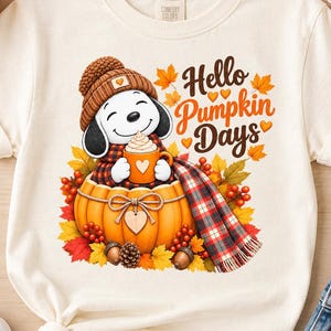 Peut inclure: Un t-shirt couleur crème avec une illustration d'un chien de dessin animé portant un bonnet en tricot marron et une écharpe à carreaux, assis dans une grosse citrouille orange en tenant une tasse. Le texte indique Hello Pumpkin Days.