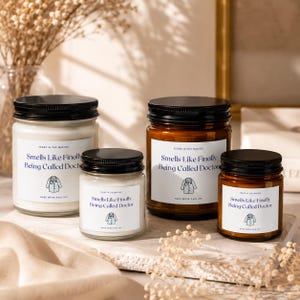 May include: Four glass candles with black lids and white labels. Two candles are in clear glass, and two are in amber glass. Each label features the text "Years in the making," "Smells Like Finally Being Called Doctor," and "Hard work paid off," with a doctor's coat icon.
