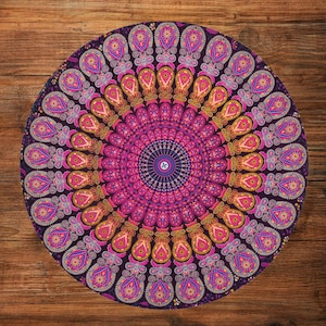 May include: A round, decorative floor cushion with a vibrant mandala design. The cushion features a symmetrical pattern in shades of purple, pink, and gold, creating a visually striking and intricate design. The cushion is placed on a wooden surface.