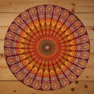 May include: A circular mandala tapestry in burgundy, featuring a detailed symmetrical design. The pattern incorporates orange, yellow, and green paisley shapes radiating outwards. The tapestry is displayed on a wooden surface.