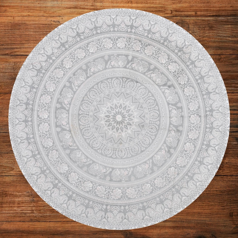 Puede incluir: Un tapiz mandala redondo, gris claro, con intrincados patrones blancos. El dise&ntilde;o presenta motivos florales y de elefantes dispuestos en c&iacute;rculos conc&eacute;ntricos, creando una est&eacute;tica detallada y sim&eacute;trica. El tapiz est&aacute; colocado sobre una superficie de madera.
