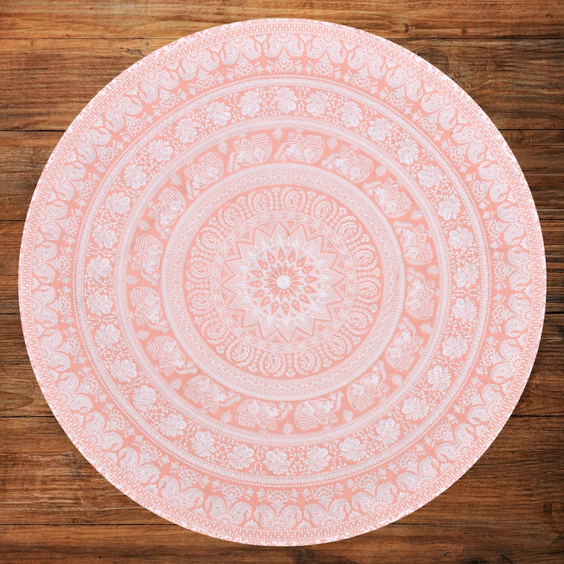 Puede incluir: Un tapiz redondo de color melocot&oacute;n con un detallado dise&ntilde;o de mandala blanco. El intrincado patr&oacute;n incluye motivos florales y de elefantes, creando una est&eacute;tica decorativa y bohemia. El tapiz est&aacute; colocado sobre una superficie de madera.