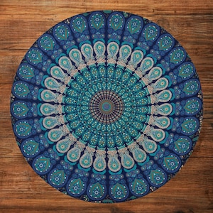 May include: Round floor cushion with a mandala design in shades of blue and teal. The cushion features a detailed pattern of teardrop shapes and circular elements, creating a symmetrical and decorative appearance. The cushion is placed on a wooden surface.