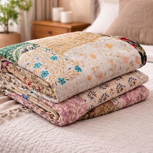 Può includere: Una pila di tre trapunte piegate con motivi floreali nei toni del verde, beige e rosa. Le trapunte sono su un copriletto bianco. La trapunta superiore ha un bordo verde e un centro beige con accenti floreali blu.