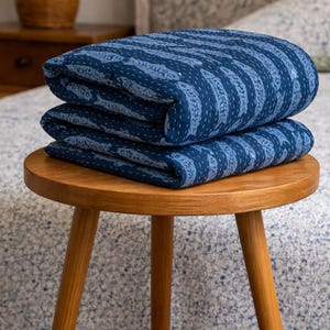May include: Two folded indigo blue kantha quilts with a striped pattern and white stitching are stacked on a small, round wooden table. The quilts are a deep blue colour with a lighter blue pattern.
