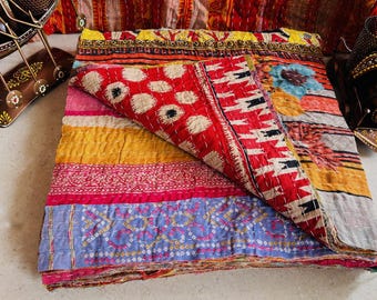 May include: A colorful, folded quilt with a patchwork design. The quilt features red, yellow, pink, and blue sections with various patterns, including polka dots and geometric shapes. The edges are stitched with white thread, and the quilt is folded to display its layered construction.