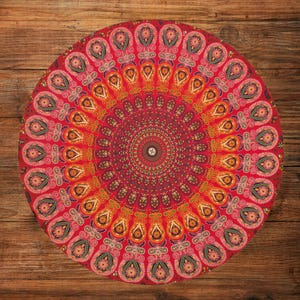 Puede incluir: Un cojín de suelo redondo rojo con un intrincado diseño de mandala. El patrón presenta una disposición simétrica de formas tipo paisley en tonos de rojo, naranja, amarillo y verde. El cojín está colocado sobre una superficie de madera.