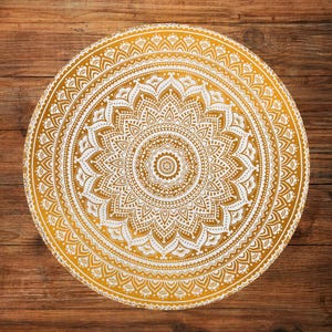 Könnte beinhalten: Ein runder, goldgelber Mandala-Entwurf mit komplizierten weißen Mustern. Das Design zeigt ein zentrales Blumenmotiv, das von konzentrischen Kreisen detaillierter, symmetrischer Elemente umgeben ist. Das Mandala ist vor einem Holzgrund gesetzt.