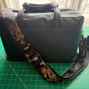 May include: Black briefcase with a decorative shoulder strap. The strap features a detailed dragon design in gold, red, and green against a black background. The briefcase has a top handle and a front zipper pocket.
