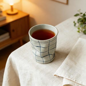 May include: A ceramic cup filled with tea, featuring a white base with a blue grid pattern. The cup sits on a white linen tablecloth, with a folded napkin to the right. A wooden side table and a small plant are in the background.