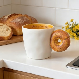 May include: A white ceramic mug with a bagel-shaped handle. The interior of the mug is yellow, and the rim has the text "One fine day." A loaf of bread and yellow flowers are in the background.