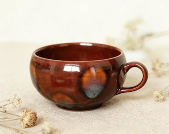 May include: A round, ceramic mug with a dark reddish-brown glaze. The mug has a small handle and a wide opening. The glaze has a glossy finish with areas of lighter brown and blue-grey hues. The mug sits on a neutral-colored surface.
