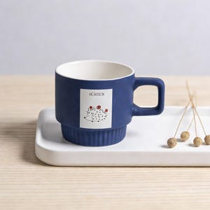 Könnte beinhalten: Eine marineblaue Keramik-Tasse mit weißem Innenraum und Henkel. Die Tasse hat ein rechteckiges Etikett mit einer Igel-Illustration und dem Wort "HERISSON". Die Tasse steht auf einem weißen Tablett.