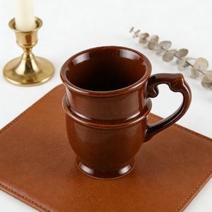 Puede incluir: Una taza de cerámica marrón con un asa decorativa. La taza tiene una base redondeada y una banda alrededor del centro. Se encuentra sobre un posavasos de cuero marrón. Un candelabro dorado y una ramita de vegetación están en el fondo.