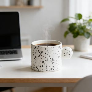 May include: A white ceramic mug with a black handle and the text "But First, Coffee" in a black, bold font. The mug is on a white surface.