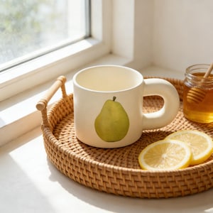 Könnte beinhalten: Eine cremefarbene Keramiktasse mit einer grünen Birnenillustration. Die Tasse steht auf einem geflochtenen Tablett mit Zitronenscheiben und einem Honigglas. Der Griff ist dick und abgerundet. Das Tablett hat einen Durchmesser von etwa 30 cm.