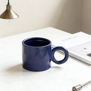 Puede incluir: Una taza de cerámica azul marino con un asa circular única. La taza cilíndrica está colocada sobre una superficie blanca. Diseño minimalista para café o té.