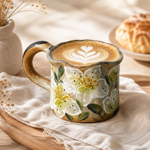 May include: A ceramic coffee mug with a floral design. The mug features white flowers with yellow centres and green leaves, set against a beige background. The mug is filled with a latte, topped with latte art. The handle is dark brown.