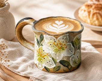 May include: A ceramic coffee mug with a floral design. The mug features white flowers with yellow centres and green leaves, set against a beige background. The mug is filled with a latte, topped with latte art. The handle is dark brown.