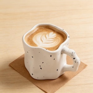 Op de afbeelding: Een witte keramische mok met een unieke, onregelmatige vorm, bespikkeld met zwarte stippen. De mok bevat een latte met een bladmotief in het schuim, rustend op een vierkante bruine papieren onderzetter. De handgreep is ook onregelmatig gevormd.