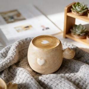 Peut inclure: Une tasse en céramique beige avec une anse en forme de cœur et un motif en forme de cœur blanc sur le devant. La tasse est remplie de café, surmontée d'un motif en mousse en forme de cœur. La tasse est posée sur une couverture en tricot grise.