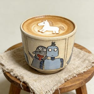以下が含まれることがあります： 白い馬のラテアートデザインが施されたベージュ色のセラミックコーヒーカップ。カップには犬とロボットの漫画イラストが描かれています。カップは、ベージュ色の布が敷かれた小さな木製のスツールの上に置かれています。