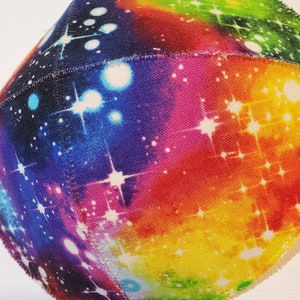 Pu&ograve; includere: Una sfera di tessuto colorata con una stampa a galassia. Il tessuto &egrave; di colore bianco con un effetto tie-dye arcobaleno e stelle bianche.