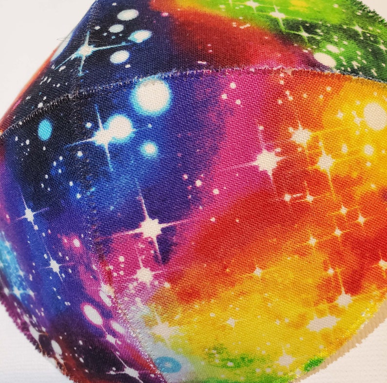 Pu&ograve; includere: Una sfera di tessuto colorata con una stampa a galassia. Il tessuto &egrave; di colore bianco con un effetto tie-dye arcobaleno e stelle bianche.