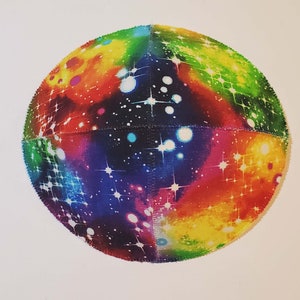 Pu&ograve; includere: Una kippah rotonda e colorata con una stampa a galassia. La kippah &egrave; realizzata in tessuto e ha uno sfondo blu scuro con stelle bianche e nebulose colorate.