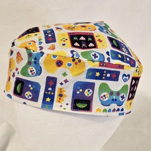May include: A white scrub cap featuring a colorful pattern of video game controllers, consoles, and other gaming-related icons in shades of blue, yellow, green, and red. The cap is designed for medical professionals or anyone in a similar field.