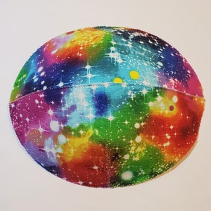 Pu&ograve; includere: Una kippa colorata con una stampa a galassia. La kippa &egrave; fatta di tessuto e ha uno sfondo bianco con un arcobaleno di colori e stelle bianche.