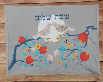 May include: A gray fabric wall hanging with a floral design featuring a branch with red pomegranates, blue leaves, and yellow flowers. Two white doves with brown spots are perched on a branch with a full moon behind them. The Hebrew text "שנה טובה" is written above the doves, which translates to "Happy New Year".