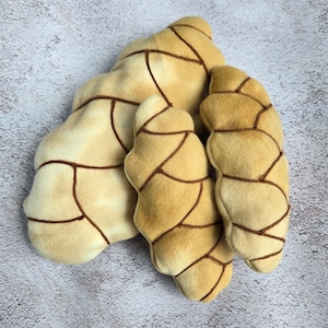 May include: Three plush pillows shaped like croissants in shades of beige and brown with brown stitching. The pillows are arranged in a pile.