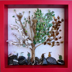 Puede incluir: Una obra de arte enmarcada que representa un árbol con cuatro estaciones, elaborada con elementos de vidrio y piedra. Aves posadas en ramas y rocas. El marco rojo proporciona un contraste visual llamativo.