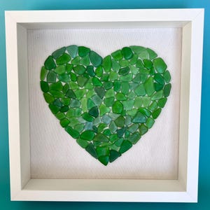 Puede incluir: Una obra de arte enmarcada que presenta un corazón compuesto por trozos de vidrio de mar verde de varios tonos. Las piezas de vidrio de mar están dispuestas sobre un fondo neutro dentro de un marco cuadrado blanco, creando un efecto de mosaico texturizado. La obra se exhibe contra una pared turquesa.