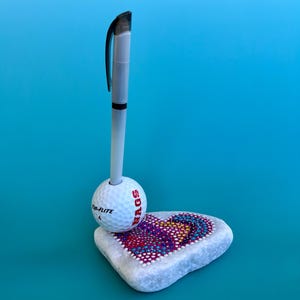Peut inclure: Un porte-stylo original avec une balle de golf et une pierre décorative en forme de cœur. La balle de golf maintient un stylo, et la pierre est peinte d'un motif de points colorés. L'ensemble est posé sur un fond turquoise.